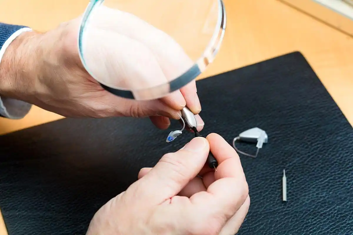 Ein Mann prüft ein Hörgerät mithilfe einer Lupe in der Hand und betrachtet die kleinen technischen Bauteile eines modernen Hörsystems in der Hörakustik.